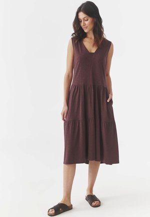 Vestido sin mangas en malva con escote en V, diseño en capas y bolsillos. Textura suave, a la altura de la rodilla, combinado con sandalias marrones tipo slip-on.