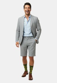 Uomo che indossa un blazer grigio chiaro e pantaloni corti, una camicia azzurra, calze verdi con motivo avocado e scarpe eleganti marroni, sorridente con una mano in tasca.
