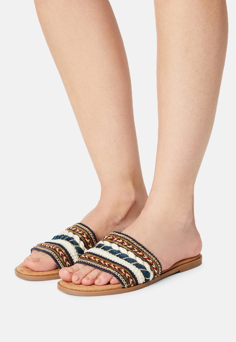 Pieds portant des sandales plates à enfiler avec des brides en tissu multicolore tressé en bleu marine, blanc, rouge et beige sur un fond neutre.