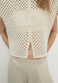 Strickoberteil in Creme mit einem Wabenmuster, runder Ausschnitt, Knopfverschluss und einem hohen niedrigen Saum, kombiniert mit hellbeigen Hosen.