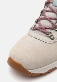 Lys beige semskesko med rosa og blå snørebånd, teksturert mesh-overdel, sølvøyer og en brun gummisåle.
