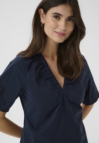 Mornarska bluza s kratkimi rokavi, V-izrezom in nabranimi detajli pri izrezu. Gladek material z sproščenim krojem in minimalističnim dizajnom.