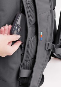 Käsi asettaa kaksoiskameroilla varustettua älypuhelinta harmaan repun vetoketjulliseen sivutaskuun, jossa on pehmustetut olkaimet ja verkkovuori.