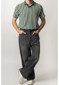 Polo rayé vert et blanc, associé à un jean noir décontracté et des baskets blanches avec des accents verts. Modèle se tenant debout avec les mains dans les poches.