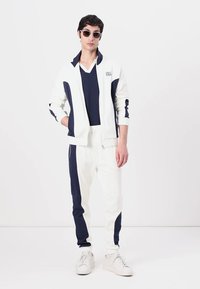 Ensemble de survêtement blanc avec des accents bleu marine, veste zippée et pantalons à poignets. Le modèle porte un t-shirt bleu marine et des lunettes de soleil, se tenant contre un fond neutre.