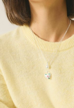 Kurze braune Haare und nackter Hals, getragen wird ein gelber, flauschiger Pullover mit mehreren silbernen Halsketten, eine mit Herz- und Blumenanhänger und eine mit Perle.