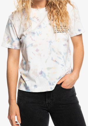 Kobieta nosząca jasnobarwną koszulkę tie-dye z krótkim rękawem z małym nadrukowanym logo na piersi oraz czarne spodnie z jedną ręką w kieszeni.