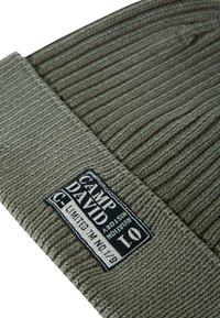 Olive green knitted beanie with ribbed texture, featuring a rectangular black label with white text: "CAMP DAVID" and additional details.