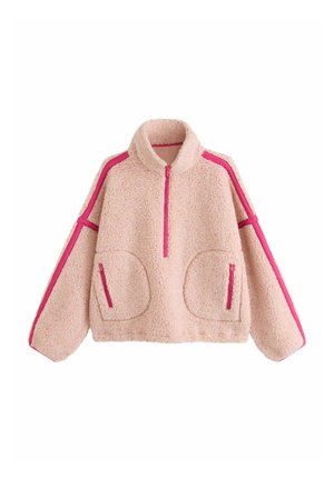 Frau mit kurzen lockigen Haaren, die eine pinke Sherpa-Fleecejacke mit magentafarbener Einfassung und passende pinke Cordshorts seitlich trägt.