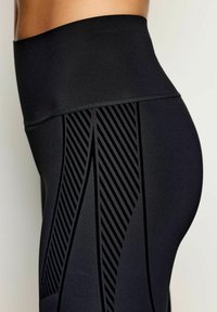 Detailní záběr osoby v černých vysokých legínách s diagonálními pruhovanými vzory po straně.