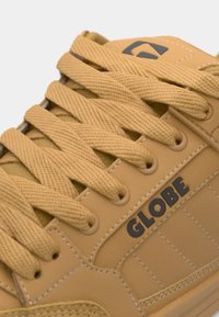 Yellow athletic shoe featuring textured upper, prominent logo, and flat laces. Design includes stitched accents and padded tongue.