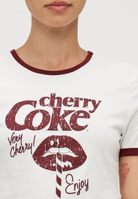 Bela majica s maroon obrobkom z vintage grafikonom "cherry Coke" in besedilom "Very Cherry!" nad stiliziranimi ustnicami in slamo.