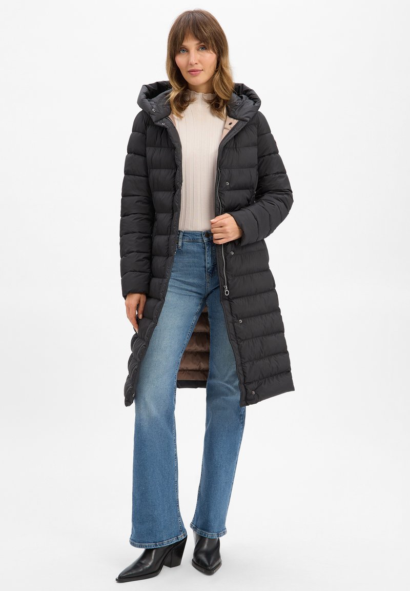 Schwarzer langer Daunenmantel mit Kapuze, gepolsterter Textur, Reißverschluss und Druckknöpfen, getragen über einem beigen Langarmoberteil und blauen Schlagjeans.