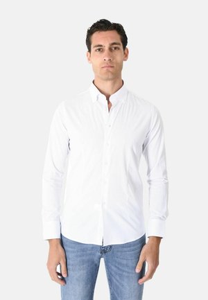 Uomo che indossa una camicia bianca semplice a maniche lunghe con bottoni e jeans blu, in piedi davanti a uno sfondo grigio chiaro.