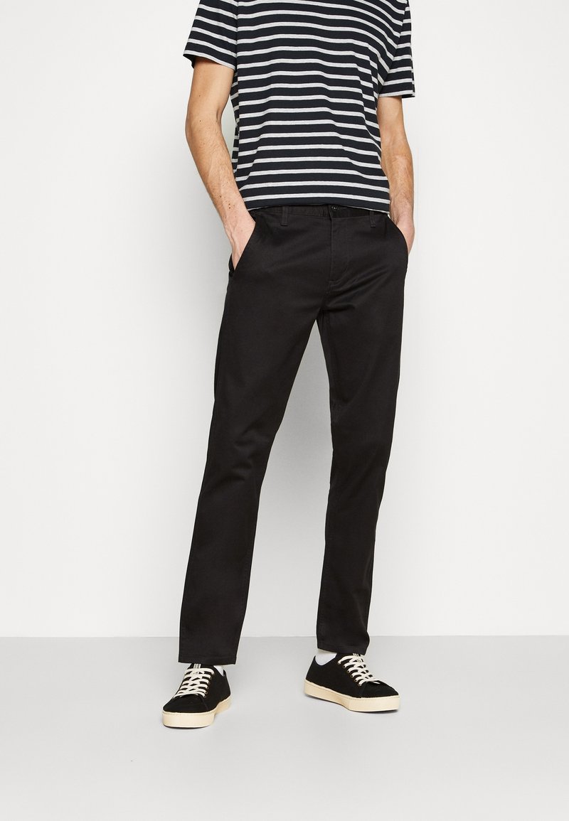 Personne portant un pantalon noir, un t-shirt à manches courtes rayé noir et blanc, et des baskets noires avec des semelles et des lacets blancs, debout les mains dans les poches.