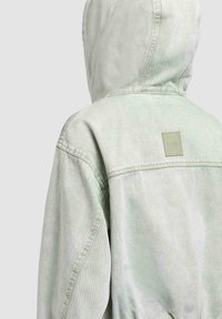 Bagsiden af en person iført en lysegrå denimjakke med hætte og en lille firkantet logo-patch øverst på ryggen.