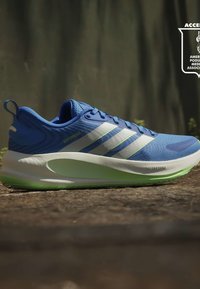 Chaussure de course Adidas bleue avec des bandes blanches et une semelle coussinée vert clair sur une surface pavée, en extérieur sous une lumière douce du jour.