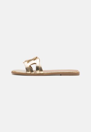 Guld flad slip-sandal med flettede brede remme og en tynd sål på hvid baggrund.