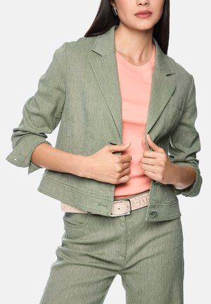 Woman wearing a light green cropped blazer and pants with a light pink shirt and matching belt, adjusting the blazer front.
