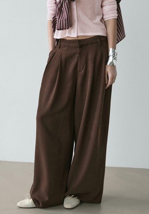 Personne portant un pantalon large marron, un cardigan court rose pâle, un bracelet manchette argenté et des chaussures à lacets beige clair, debout devant un fond uni.