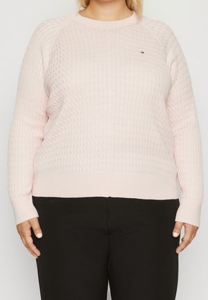 Femme portant un pull en maille texturée rose clair et un pantalon noir, debout devant un fond clair uni.