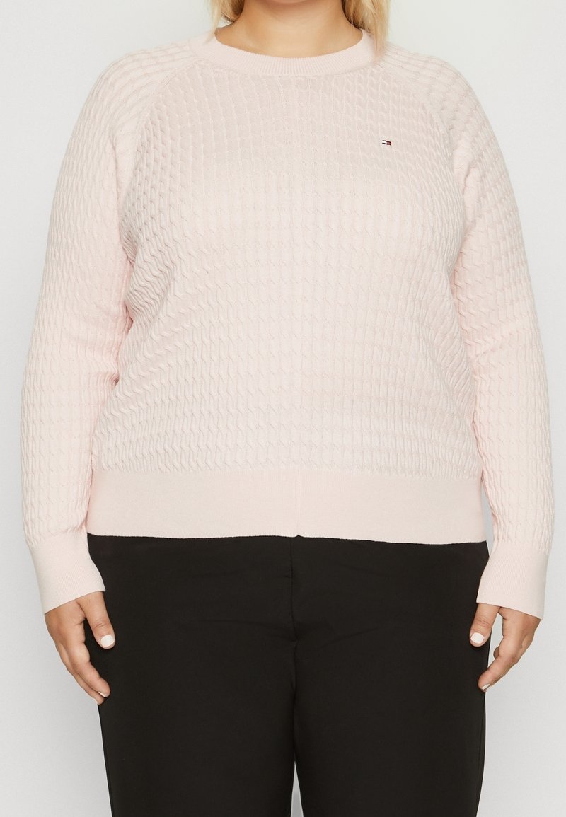 Femme portant un pull en maille texturée rose clair et un pantalon noir, debout devant un fond clair uni.