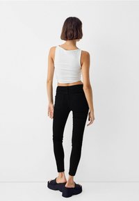 Mujer con cabello corto castaño se encuentra frente a una pared blanca, vistiendo un top de tirantes blanco, jeans ajustados negros y sandalias de plataforma oscuras.