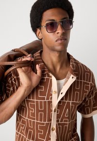 Brown patterned short-sleeve shirt with beige accents and buttons, worn with a white undershirt. Model holds a brown leather bag. Dark sunglasses.