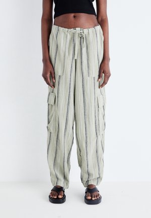 Person trägt beige, locker sitzende, hochtaillierte Cargohosen mit vertikalen Streifen, Kordelzug in der Taille, aufgesetzte Taschen, schwarzes Croptop und schwarze Sandalen.
