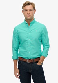 Turquoise overhemd met lange mouwen, voorzien van een kraag en een klein logo. Gepaard met donkere jeans en een leren bruine riem.