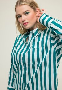 Camisa a rayas en azul verdoso y azul claro, hecha de tejido suave. Presenta un cierre frontal con botones, cuello de punta y dos bolsillos en el pecho.