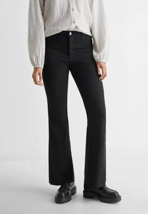 Czarne dżinsy dzwonowe z wysokim stanem, z surowym wykończeniem i subtelnymi detalami szwów. Połączone z czarnymi butami na platformie i teksturowaną białą bluzką.