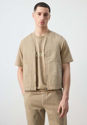 Joven con camisa beige de manga corta, chaleco a juego y pantalones con detalles de salpicaduras de pintura, de pie con las manos en los bolsillos.