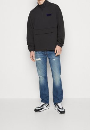 Sudadera negra con cremallera, de manga larga, con un bolsillo frontal y texto bordado. Combinada con jeans desgastados de color azul claro y zapatillas negras y blancas.