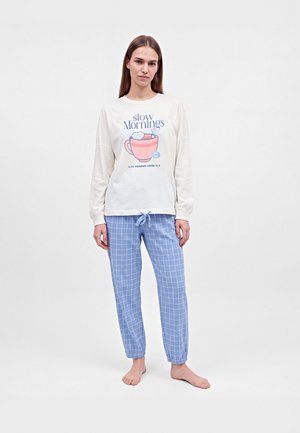 Une femme porte un sweat-shirt blanc avec l’inscription « Slow Mornings » et un pantalon de pyjama bleu à carreaux. Elle se tient pieds nus devant un fond de studio uni.