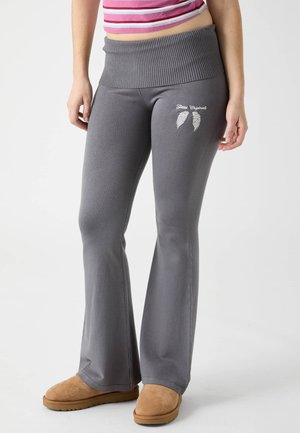 Donna che indossa pantaloni a zampa di elefante in maglia grigia con logo a forma di ala sulla coscia, top corto a righe rosa e scarpe slip-on color sabbia, in piedi su sfondo bianco.