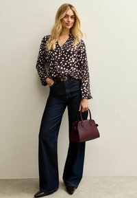 Donker gemêleerd blouse met lange mouwen, high-waisted blauwe jeans en een kleine bordeauxrode handtas. Model staat tegen een neutrale achtergrond.