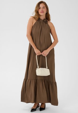 Femme en robe longue marron sans manches avec un détail fleuri au cou, tenant un sac tressé crème, portant des talons noirs, debout.