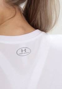 Bílé sportovní tričko s kulatým výstřihem, s malým šedým logem Under Armour na zádech. Měkká, hladká textura látky.