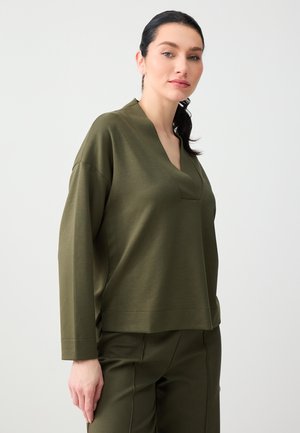 Donna con capelli scuri raccolti, indossa un top verde oliva a maniche lunghe con scollo a V e pantaloni coordinati, in piedi contro uno sfondo chiaro e uniforme.