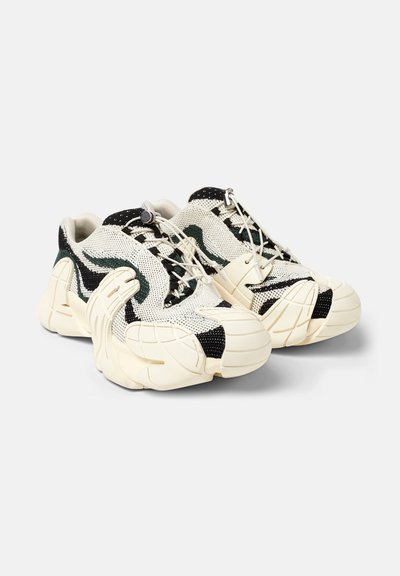 Zapatillas de malla con una base crema, acentos en negro y verde, superficies texturizadas y suelas blancas gruesas. Cuenta con un sistema de cordones único.