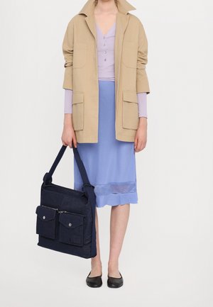 Žena v béžové bundě, levandulovém topu, modré sukni s síťovým detailem, černých balerínách, držící velkou tmavě modrou kabelku přes rameno s předními kapsami.