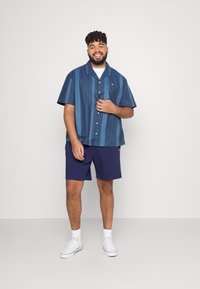 Kortærmet skjorte med knapper og lodrette blå striber, parret med mørkeblå shorts. Har et lille logo på brystlommen.