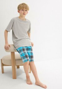 T-shirt en coton gris, coupe décontractée, associé à un short à carreaux bleu, noir et blanc. Assis sur une chaise claire. Pieds nus.