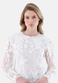 Weiße Langarmbluse mit kunstvoller floralem Stickerei, transparenter Textur und rundem Ausschnitt. Verfügt über detaillierte Muster an den Ärmeln und auf der Vorderseite.