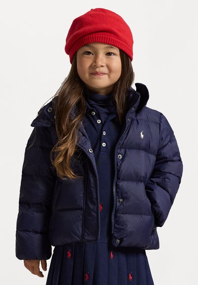 Navy puffer jacket with snap buttons, featuring a hood and a small white logo. Red knit hat. Underneath, a navy dress with red accents.