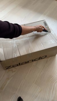Pahvilaatikko, jossa on Zalandon brändäys. Suljettu kirkkaalla teipillä. Käsi pitää askarteluveistä, joka leikkaa teippiä laatikosta. Vaalea puulattia.