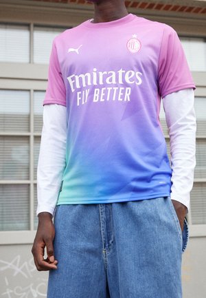 Mann trägt ein AC Milan Fußballtrikot mit Verlauf von Pink zu Blau mit Emirates Fly Better Logo, darunter weiße langärmlige Unterziehshirt und blaue Jeans.