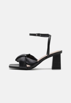 Schwarze Leder-Sandalette mit offenem Zehenbereich, gepolstertem verdrehtem Vorderriemen, Knöchelriemen mit Schnalle und Blockabsatz auf einfachem hellem Hintergrund.