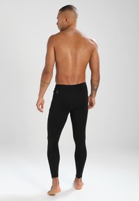 Fekete leggings egy személy hátulján láthatóak. Az anyag simának tűnik, a fazon pedig testhezálló, a derékpánt közelében pedig egy lehetséges logó is látható.